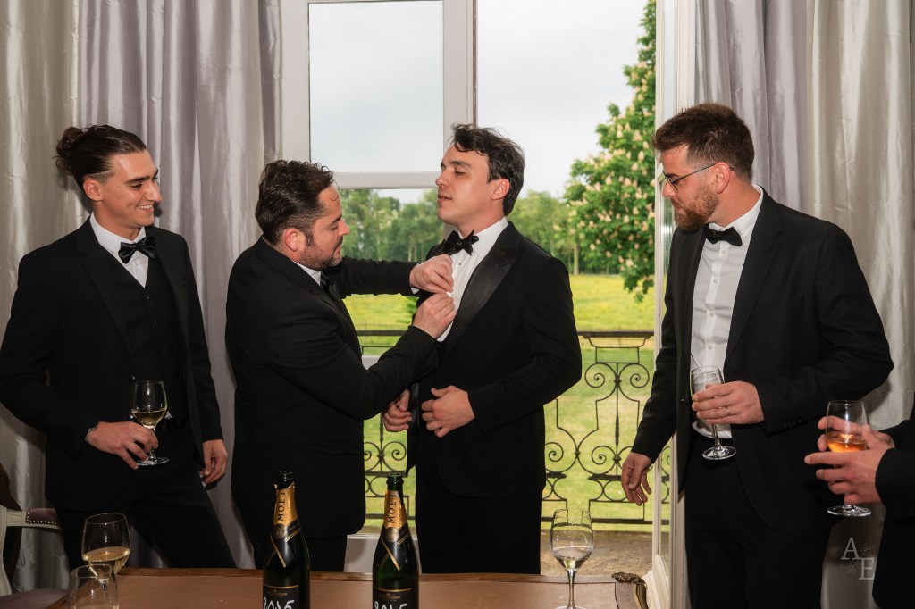 Groomsmen helping groom with bow tie in getting ready suite in France with champagne and countryside view